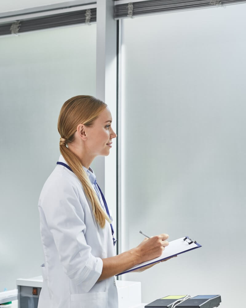 Side Image Side View Waist Up Portrait Of Beautiful Young Lady In White Lab Coat Doing Paperwork While Standing In Medical Office.